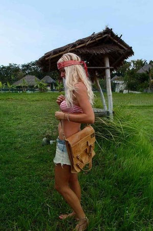 A person standing on grassy ground next to a traditional structure, carrying a brown foldover tote bag with leather straps.