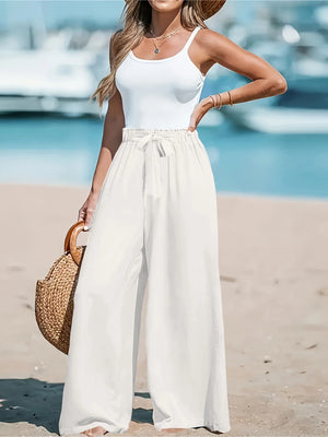 Woman in white tank top and wide-leg pants standing on a beach with a straw bag.