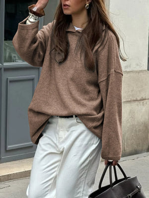 Person wearing a brown sweater and white pants, holding a black handbag on a city street.