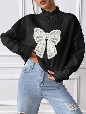Person wearing a black sweater with a large white bow design, standing against a white wall.