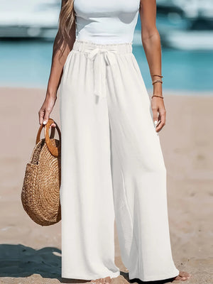 Person wearing white pants and a light blue top on a beach, holding a woven bag.