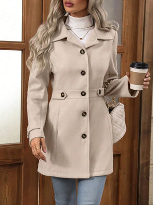 Woman wearing a beige coat with black buttons, holding a coffee cup, standing in front of a wooden door.