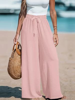 Person wearing pink wide-leg pants and a white top on a beach, holding a straw bag.