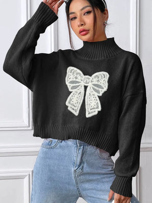 Person wearing a black sweater with a white bow design against a white wall.