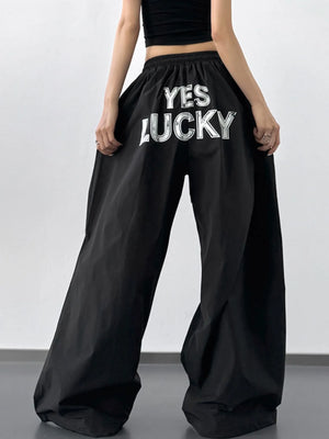 Black wide-leg pants with 'YES LUCKY' text worn by a person on a plain background
