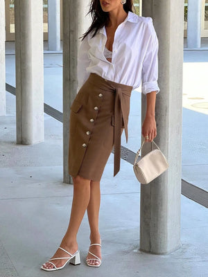 Woman wearing a white shirt and brown skirt with buttons, holding a beige handbag.