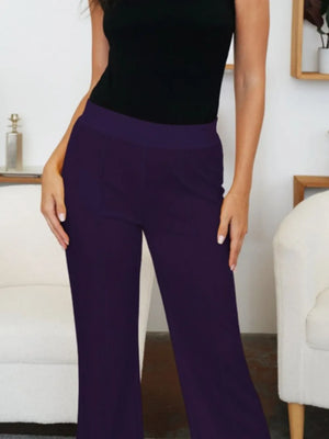 Person wearing purple pants and a black top in a room with a white chair and a shelf.