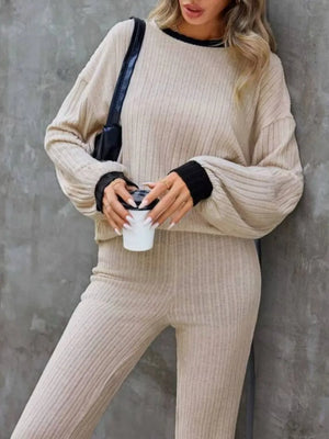 Person wearing a beige ribbed sweater and pants holding a white cup against a gray wall.