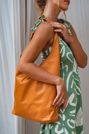 A person wearing a green floral dress is holding an orange slouchy leather shoulder bag with a leather wrapped shoulder strap.