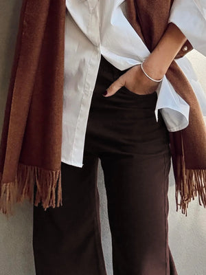 Person wearing a white shirt, brown pants, and a brown scarf against a neutral background