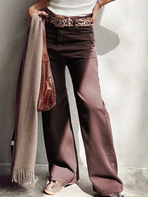 Person wearing wide-leg brown pants with a white shirt and patterned belt, holding a beige scarf.