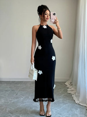Woman wearing a black halter neck dress with white floral patterns, standing in a room with light-colored curtains.
