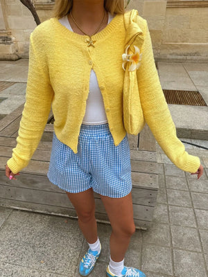 Person wearing a yellow cardigan, white top, blue checkered shorts, and blue sneakers on a stone pavement.