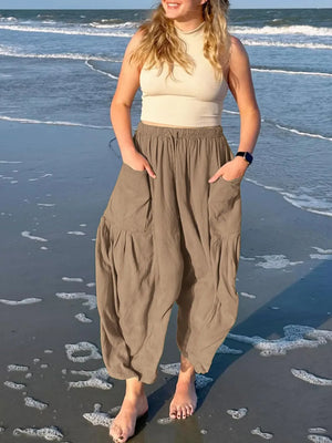 Woman standing on a beach wearing a beige sleeveless top and brown harem pants.