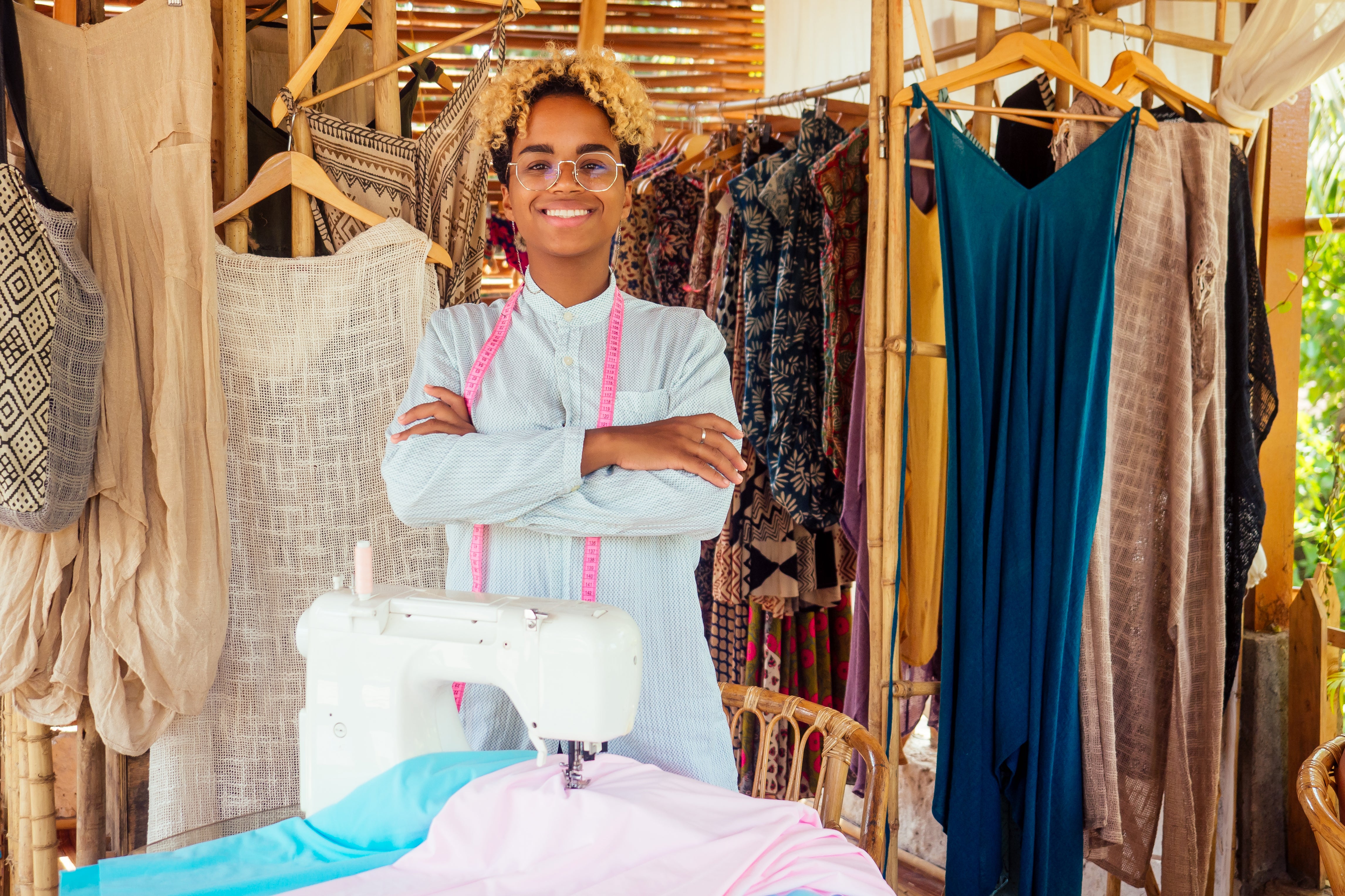 Lady standing in front of a rack of clothes with a tape measure around her neck with a sowing machine in front of her on a table