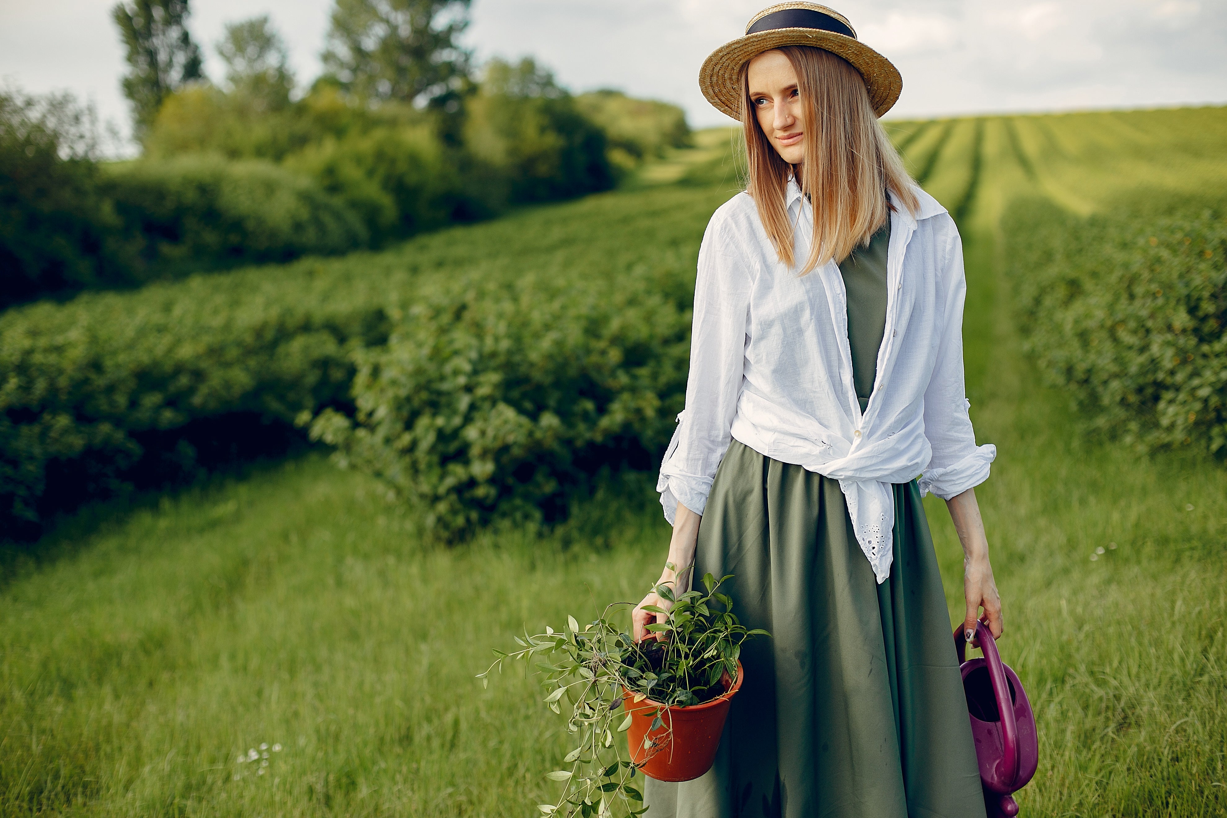 Woman standing in a green field with a green dress on with a white shirt over it and a plant in one hand a handbag in the other hand