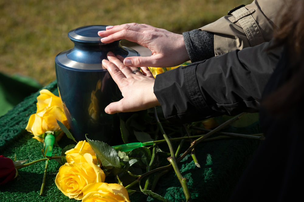 A picture of hands on an urn with yellow flowers around it.