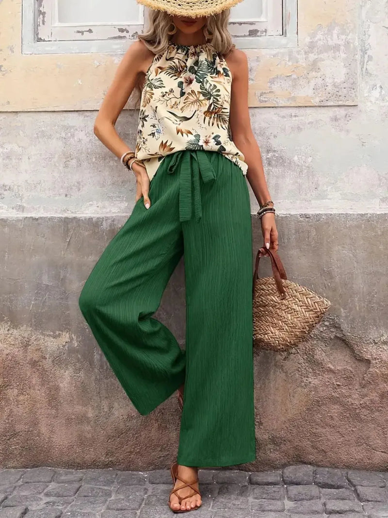 Woman wearing a floral top and green pants against a textured wall.