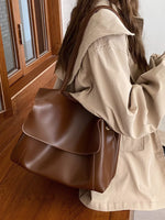 Person wearing a beige coat holding a brown leather bag indoors.