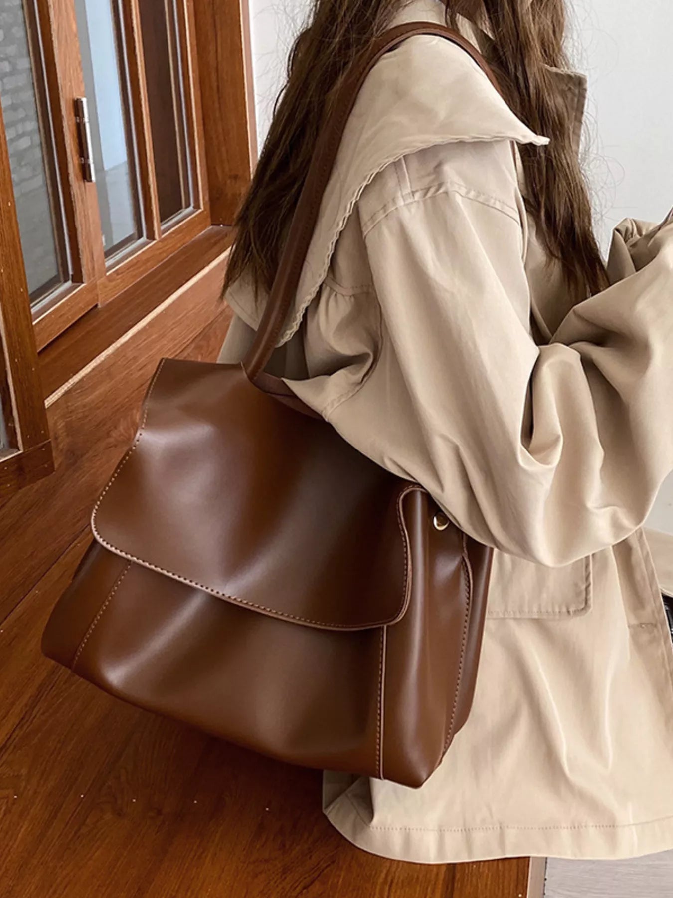 Person wearing a beige coat holding a brown leather bag indoors.