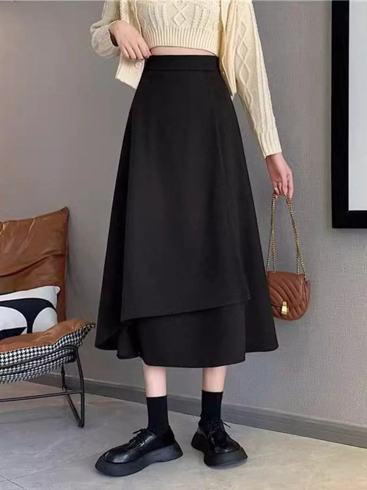Person wearing a black skirt with a beige sweater and black shoes in an indoor setting.