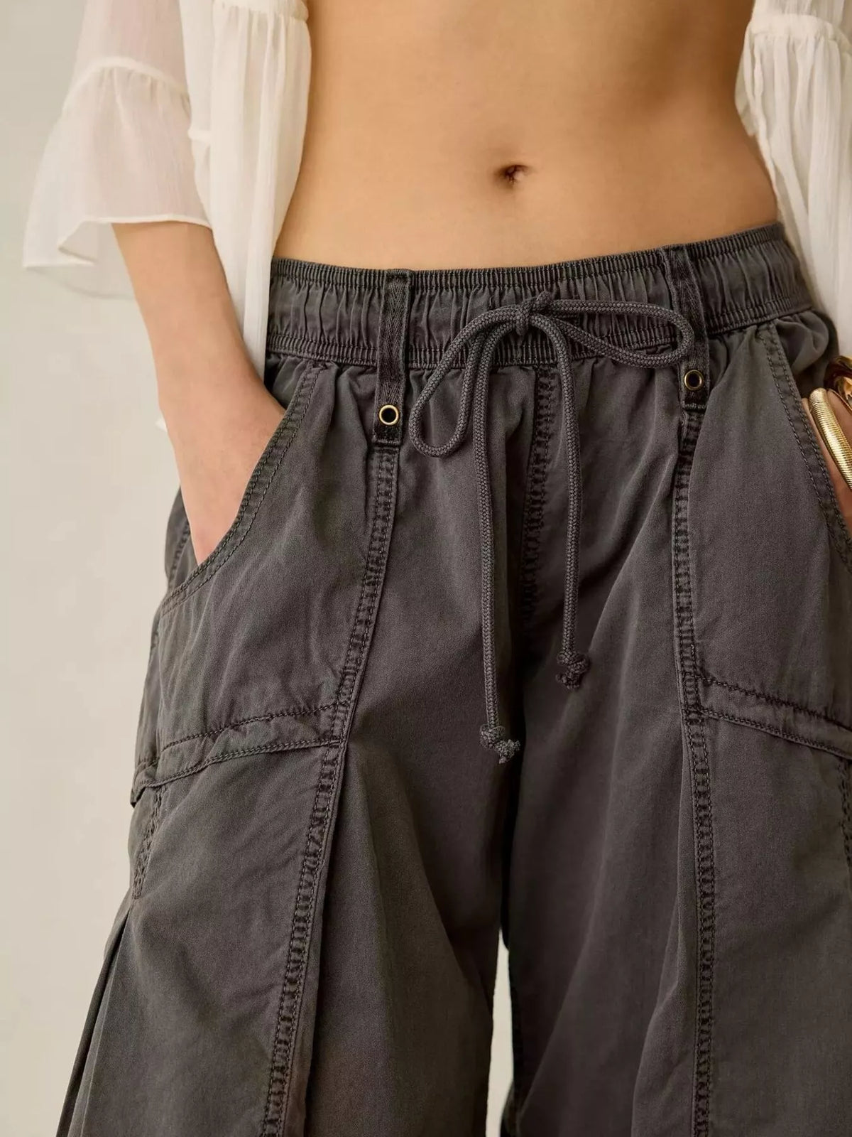 Close-up of a person wearing dark charcoal gray wide-leg cargo pants with an elastic drawstring waist, side pockets, and contrast stitching. A white ruffled blouse is partially visible.