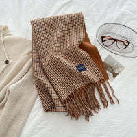 Plaid scarf with fringes on a white surface next to a beige cardigan and glasses.