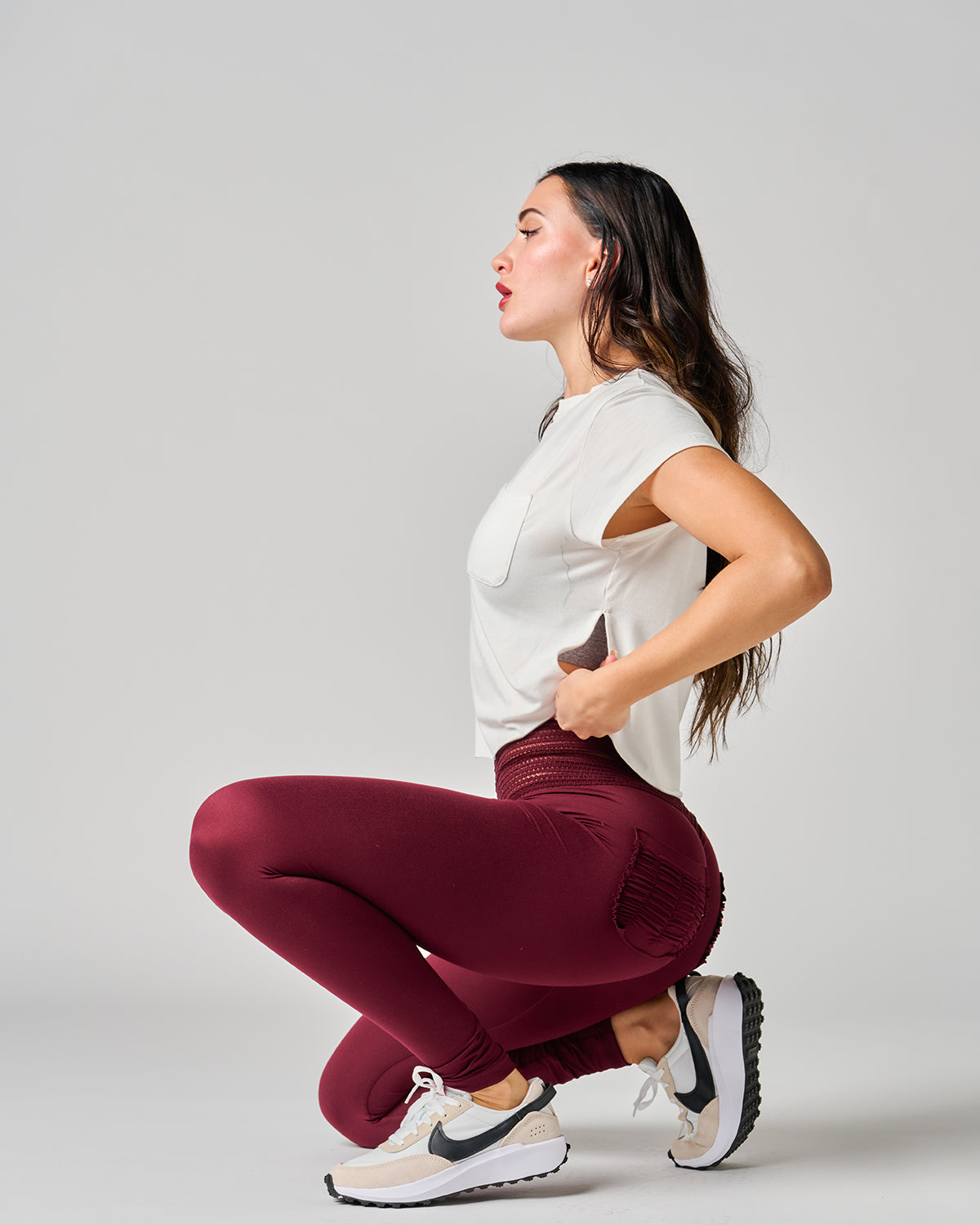 Woman in white shirt and maroon leggings squatting on a light gray background