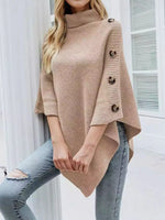 Woman wearing a beige poncho with button details over light blue jeans indoors.