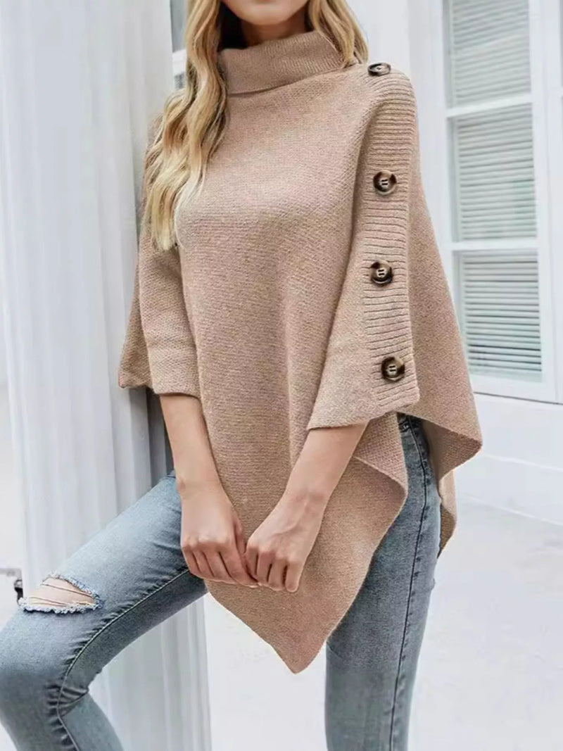 Woman wearing a beige poncho with button details over light blue jeans indoors.