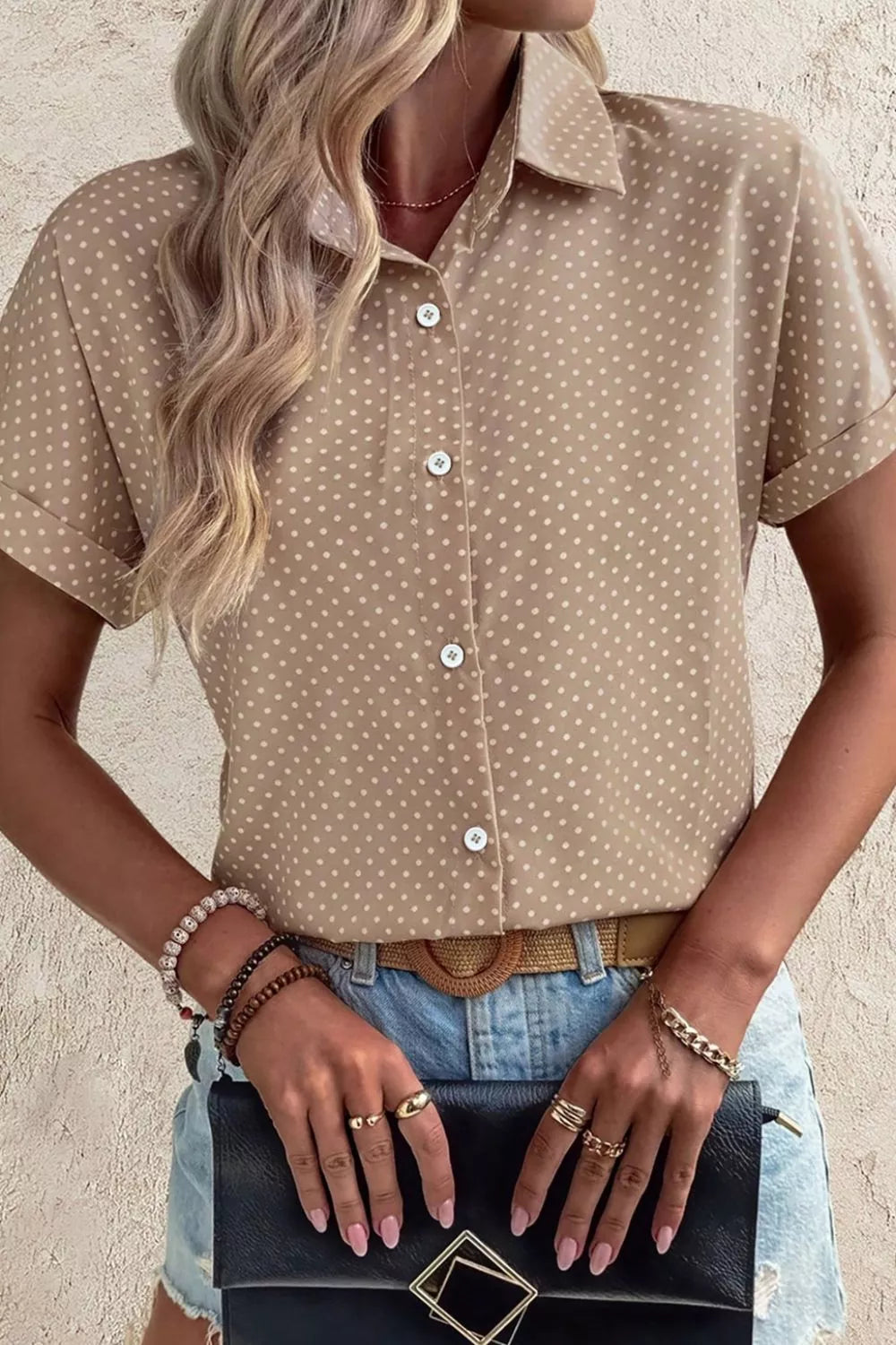 Woman wearing a beige polka dot shirt with denim shorts, holding a black clutch.