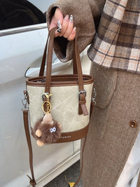 Person holding a beige handbag with brown accents and a fluffy keychain, wearing a plaid jacket and brown coat.