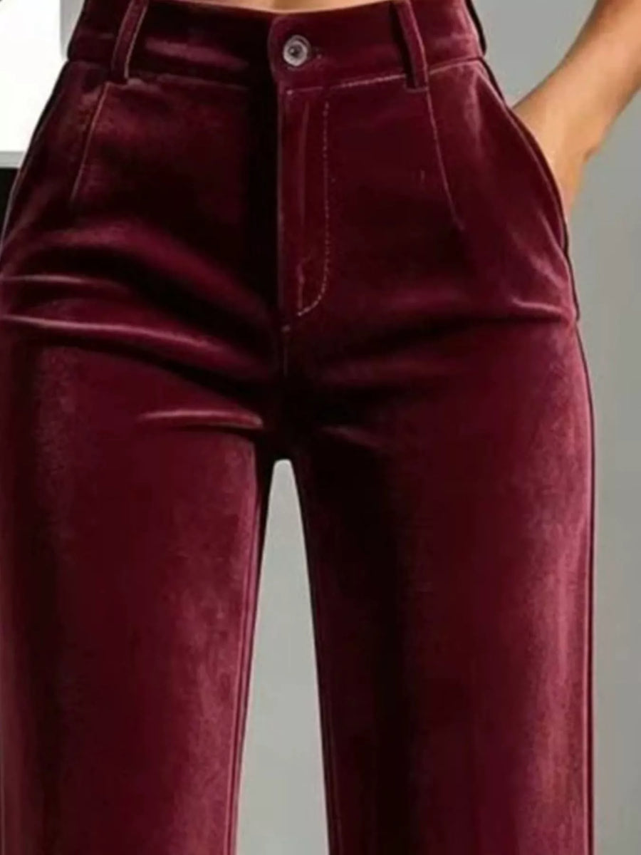 A close-up of a person wearing high-waisted burgundy velvet pants, showing the front waistband, button, fly, and front pockets.