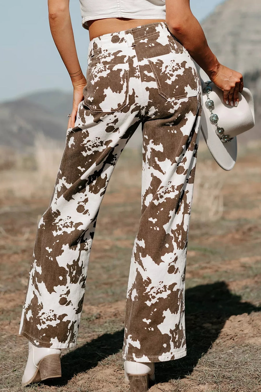 A person outdoors wearing high-waisted, wide-leg pants with a brown and white cow-print pattern, holding a white western-style hat.