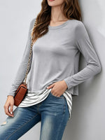 A woman wearing a light gray long-sleeve crewneck top, blue jeans, and a brown crossbody bag with a gold chain strap, leaning against a light gray wall.