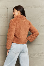 Person wearing a brown fuzzy jacket and light blue jeans against a neutral wall.