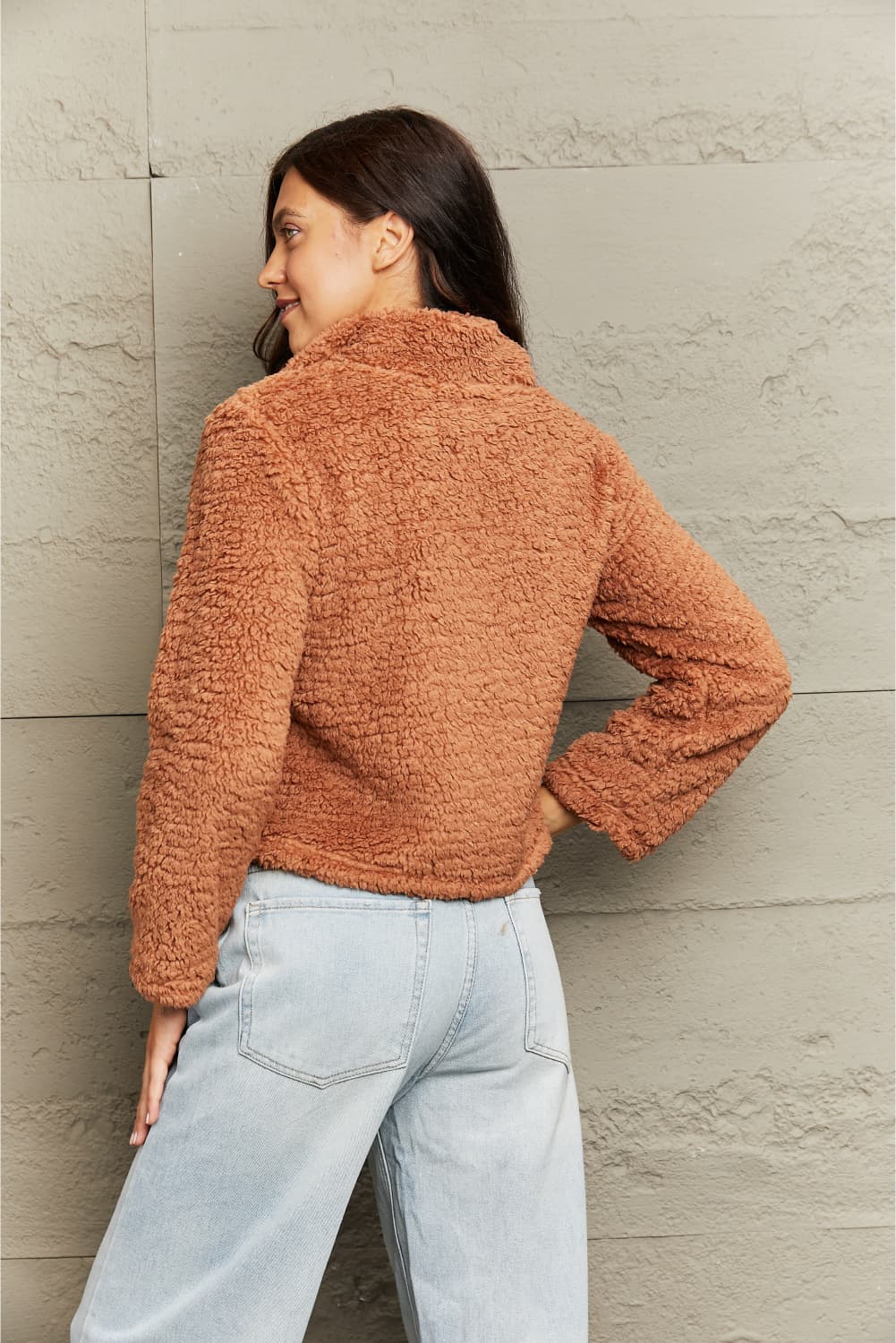 Person wearing a brown fuzzy jacket and light blue jeans against a neutral wall.