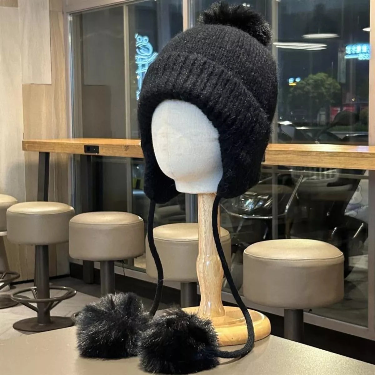 Black knitted hat with pom-poms on a mannequin head in a cafe setting