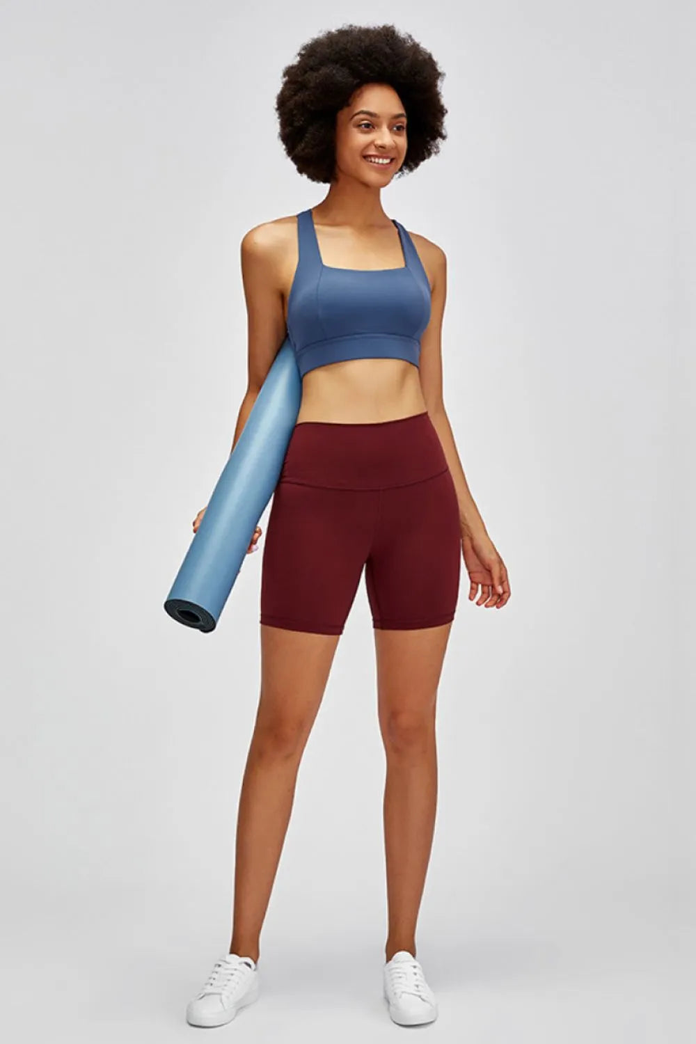 Woman holding a blue yoga mat, wearing a blue sports bra and maroon shorts on a light gray background