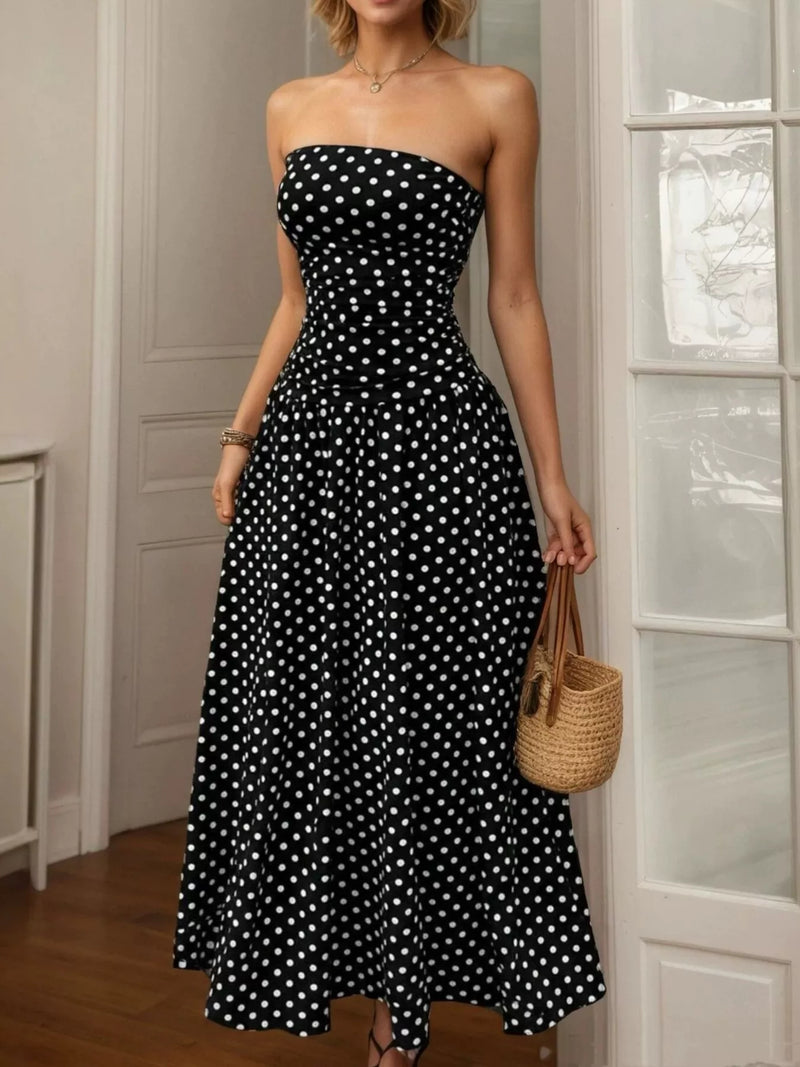 Woman wearing a black strapless dress with white polka dots, holding a woven bag indoors.
