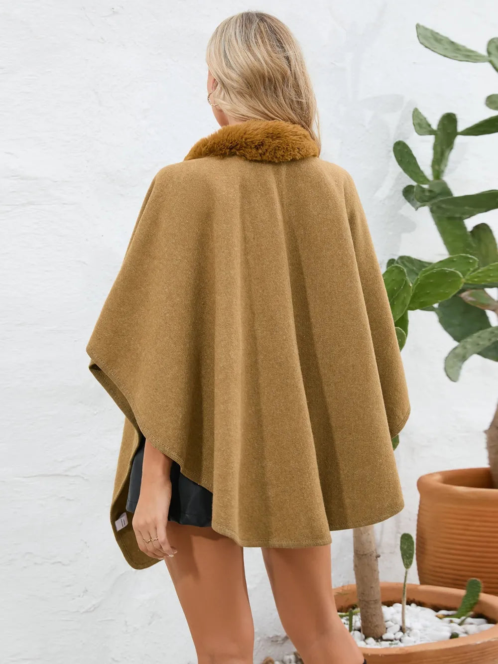 Person wearing a brown cape with a fur collar in front of a white wall and potted plants.