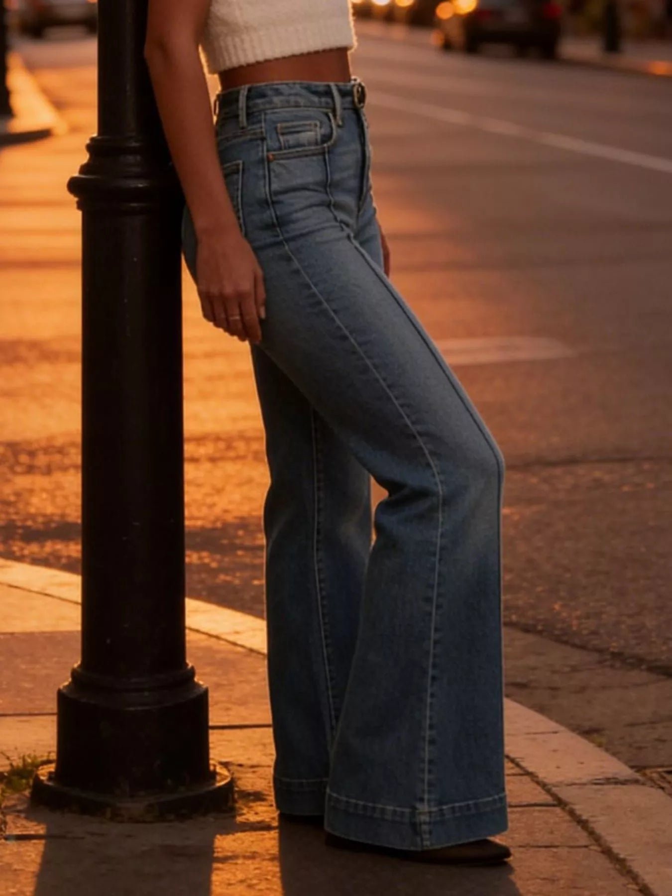 Person wearing blue jeans standing on a sidewalk with a street lamp in the foreground.