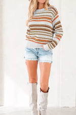 A woman wearing a long-sleeve knit sweater with horizontal stripes in orange, brown, olive green, and white, paired with light-wash denim shorts and off-white knee-high boots.