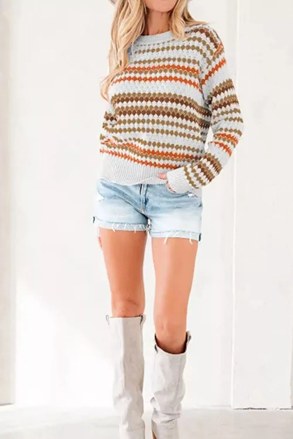 A woman wearing a long-sleeve knit sweater with horizontal stripes in orange, brown, olive green, and white, paired with light-wash denim shorts and off-white knee-high boots.