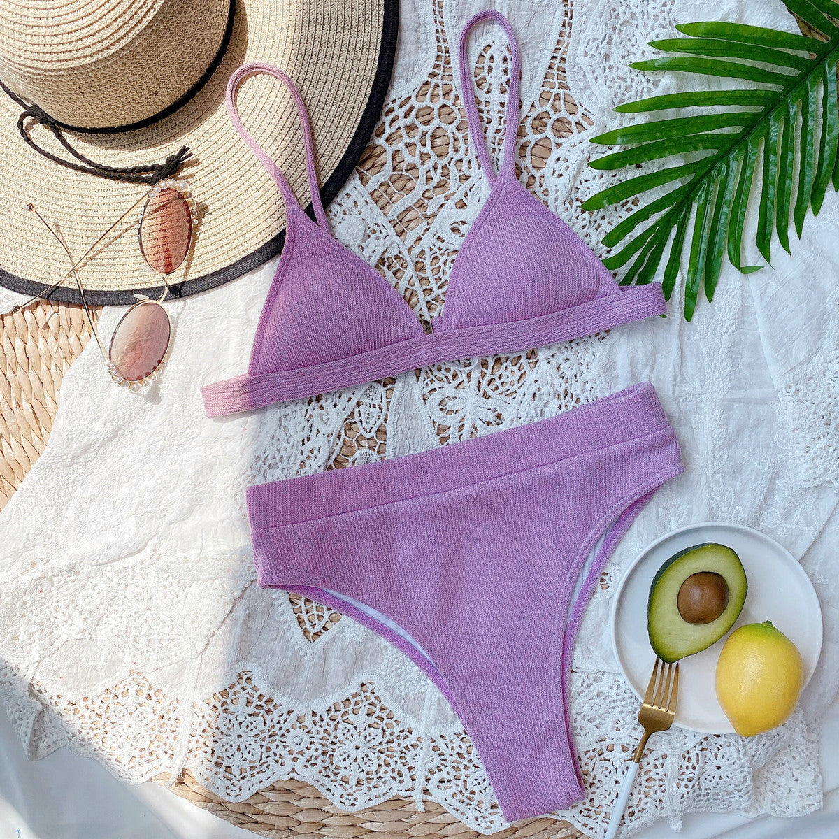 Purple bikini set on a white lace surface with a hat, sunglasses, and avocado on a plate.