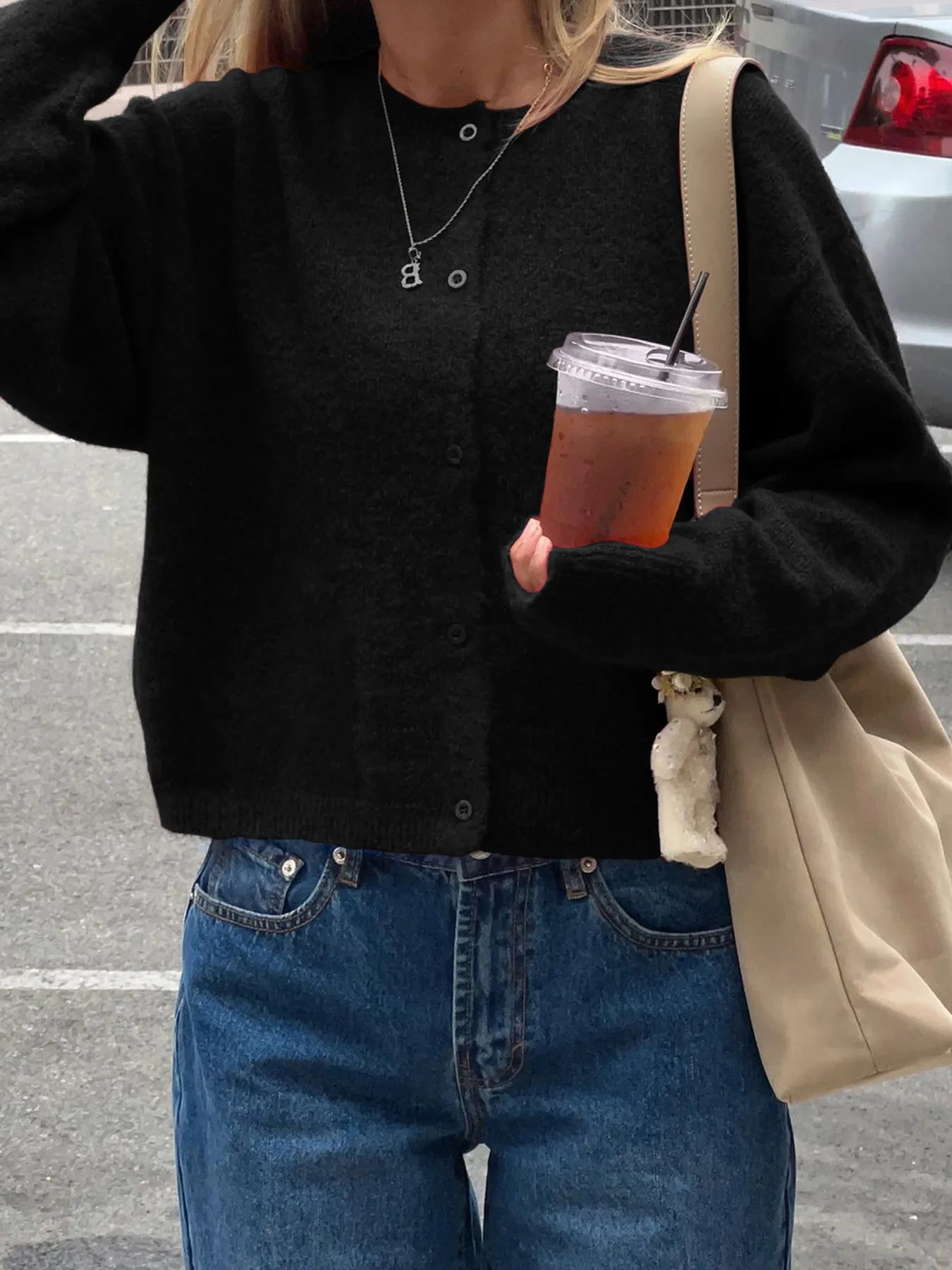 Person wearing a black cardigan and blue jeans, holding a drink and a beige bag.