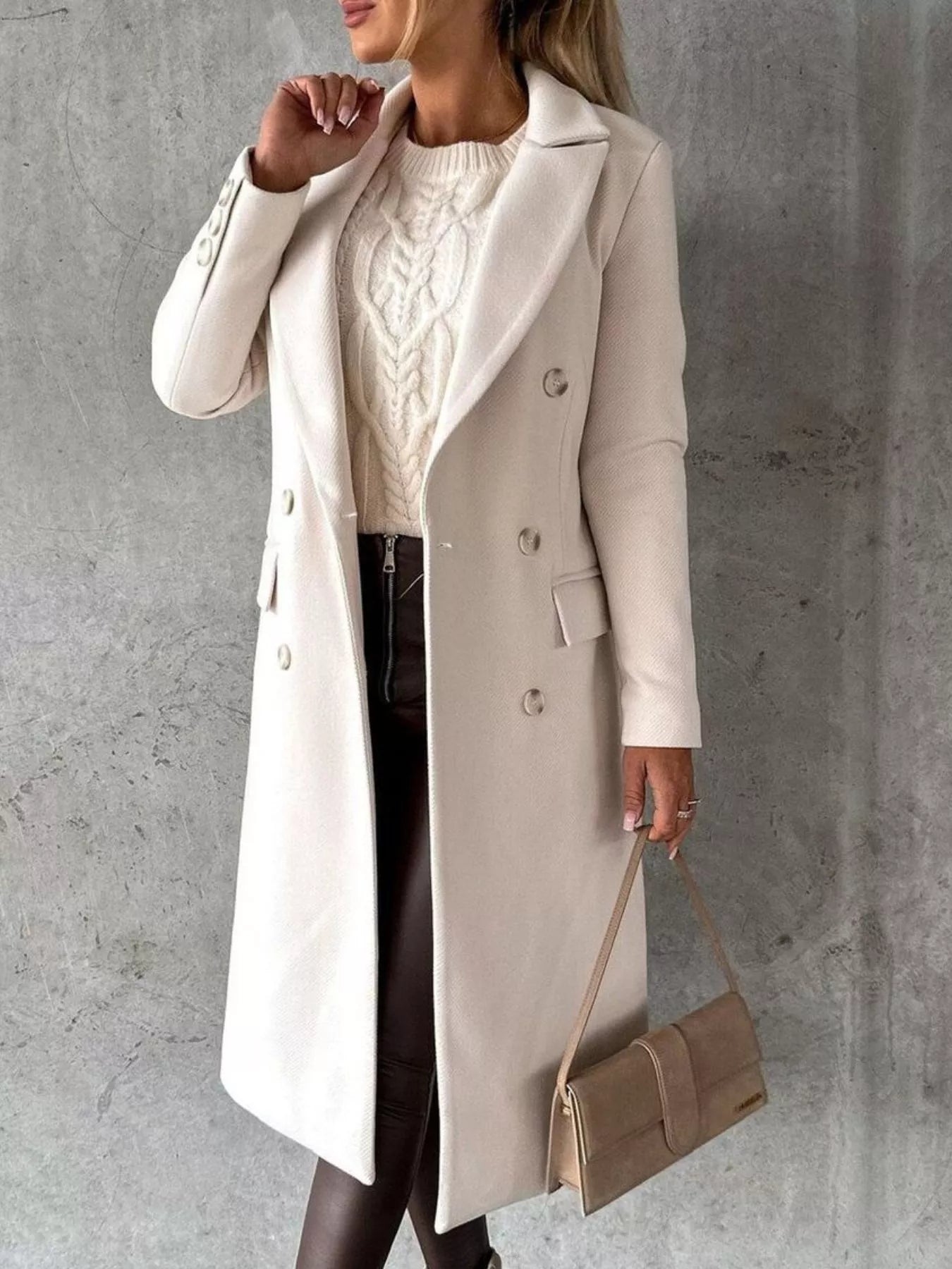 Woman wearing a long beige coat and holding a beige handbag against a gray background