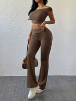 Woman wearing a brown outfit with high-waisted pants and a matching top, holding a brown handbag against a white wall.