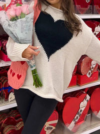 Person wearing a white sweater with a black heart design, holding flowers in a store setting.