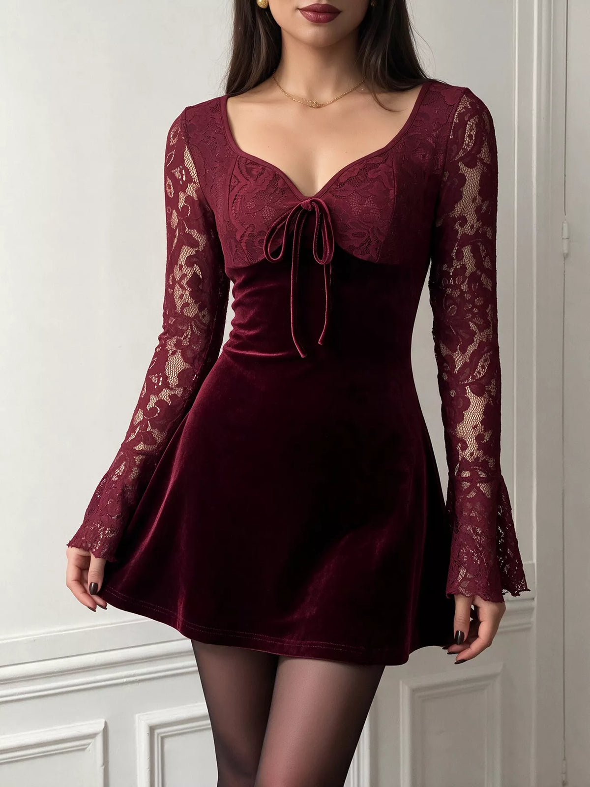 A woman wearing a burgundy velvet mini dress with long lace sleeves, a sweetheart neckline, and a front tie detail, paired with sheer black tights.